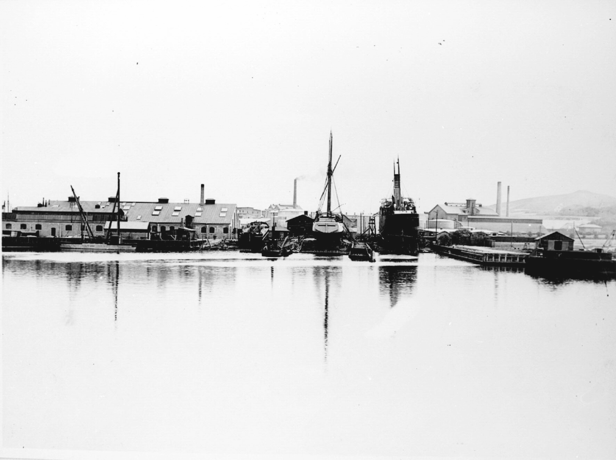 Historic shipyard with vessels docked, industrial buildings, and calm water reflection.