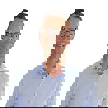 Hombre sonriente con gafas y una camisa azul claro sobre un fondo blanco.
