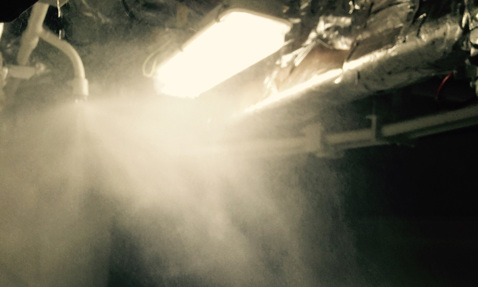 Bright industrial light illuminating mist from overhead pipes in a dimly lit area.