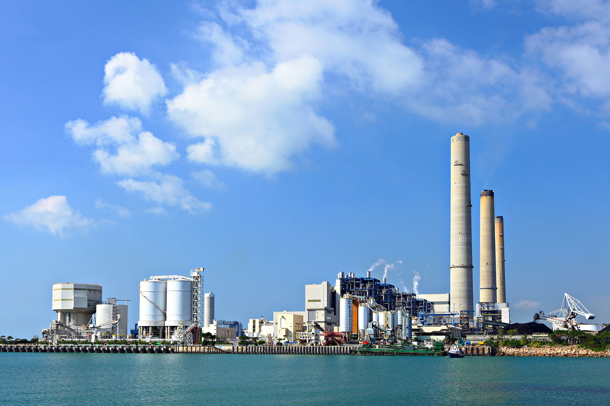 Industrial power plant with tall smokestacks near water under a clear blue sky.