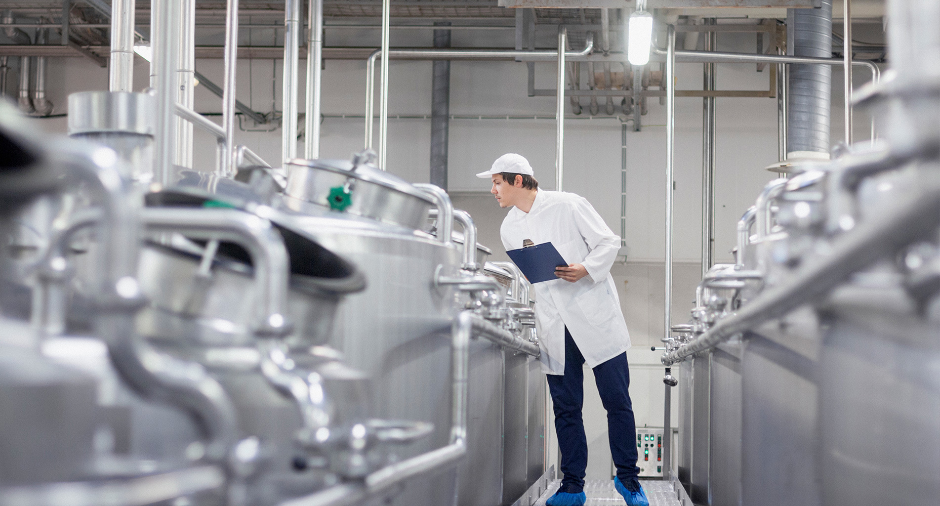 Worker inspecting stainless steel tanks in an industrial facility, holding a clipboard.