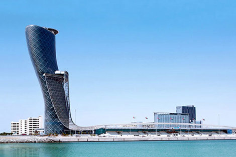 Modern, leaning skyscraper against a clear blue sky, reflected in calm water.