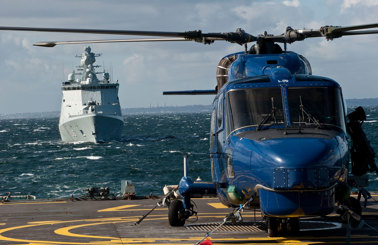 Blue helicopter on a ship's deck with a naval ship approaching.