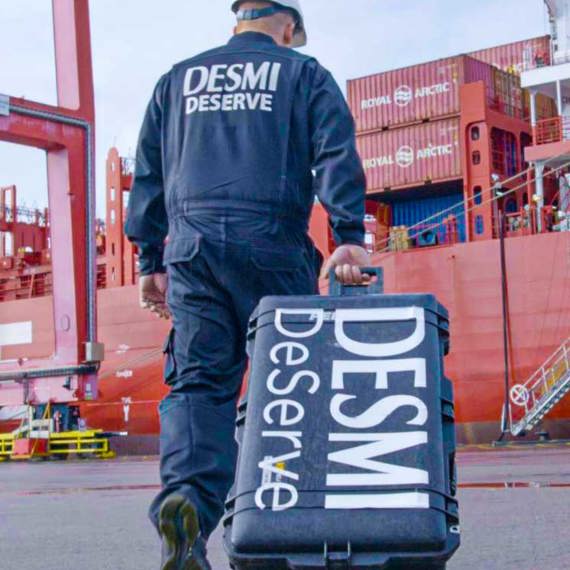 Worker in DESMI uniform carrying equipment case at a shipping port.