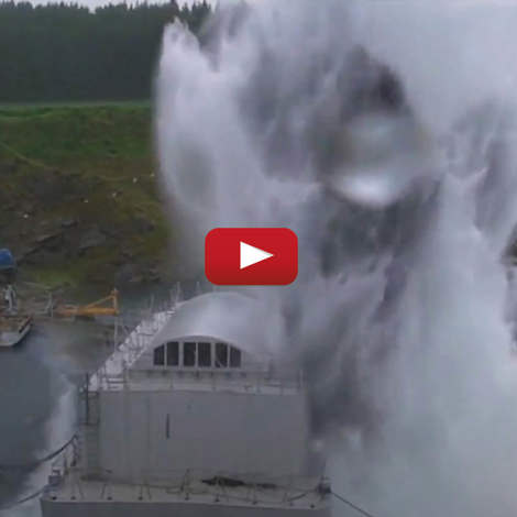 Large explosion in water near industrial structure with forested background.