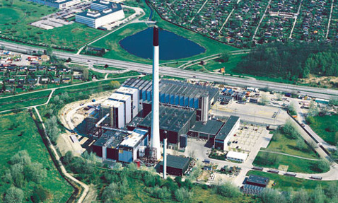 Aerial view of a large industrial plant with a tall smokestack and surrounding greenery.