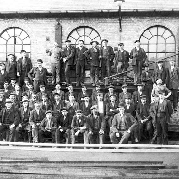 Large group of men in work attire posed outside a brick building.
