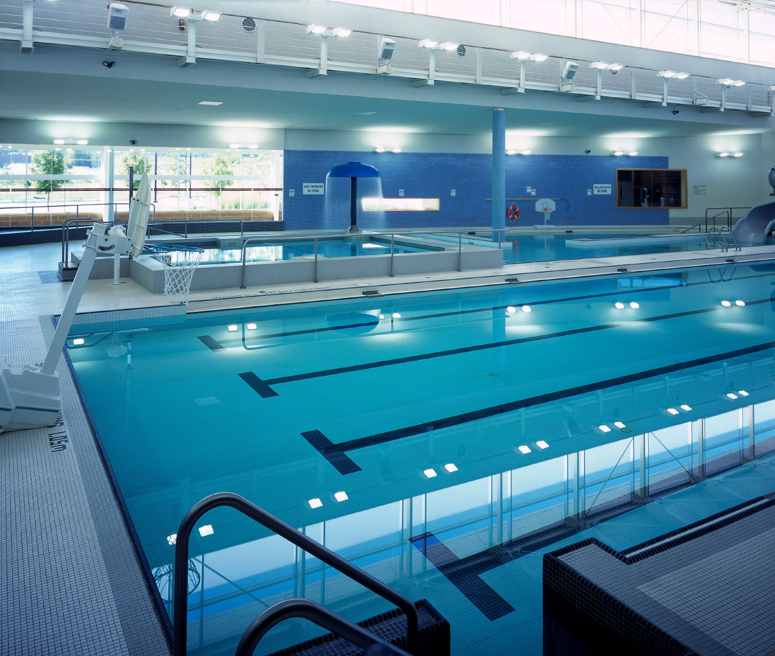 Indoor swimming pool with diving boards, basketball hoop, and bright overhead lighting.