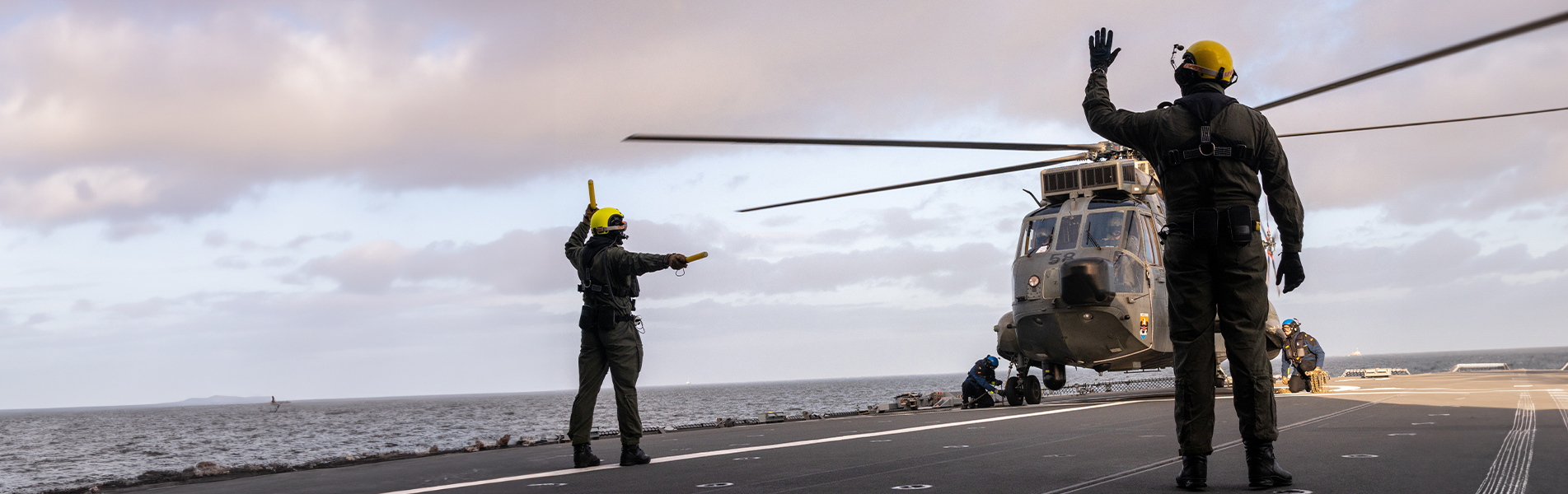 Flight deck crew guiding helicopter landing on aircraft carrier at sea.
