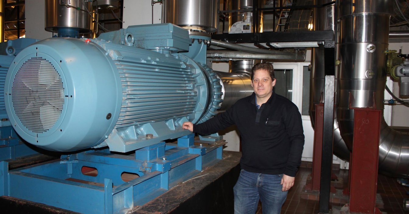 Man standing beside large blue industrial machine in a mechanical room.