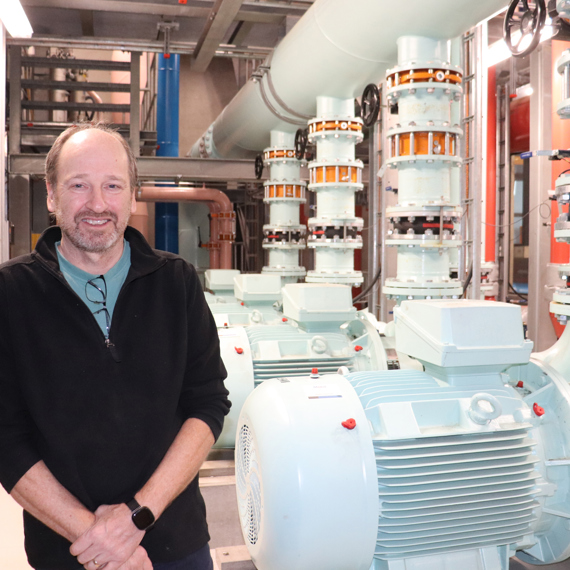 Man standing in an industrial facility with large machinery and pipes in the background.