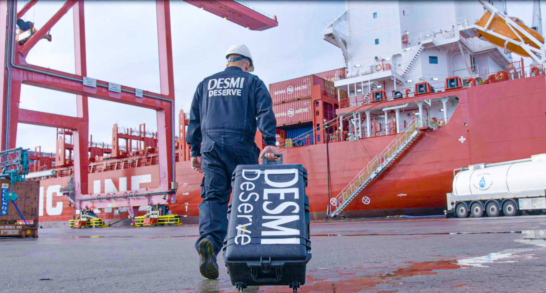 Worker in DESMI uniform pulling a suitcase near a large cargo ship.